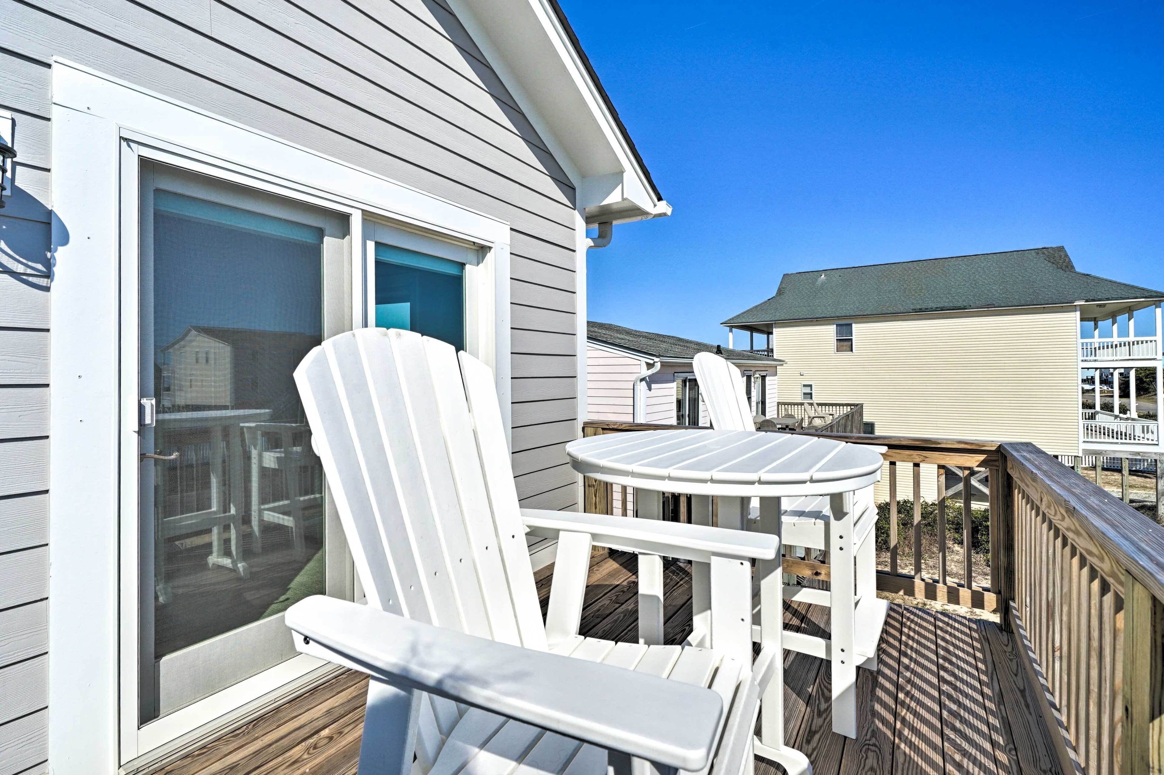 A deck with white Adirondack chairs and a small round table, overlooking a wooden deck and a neighboring house.
