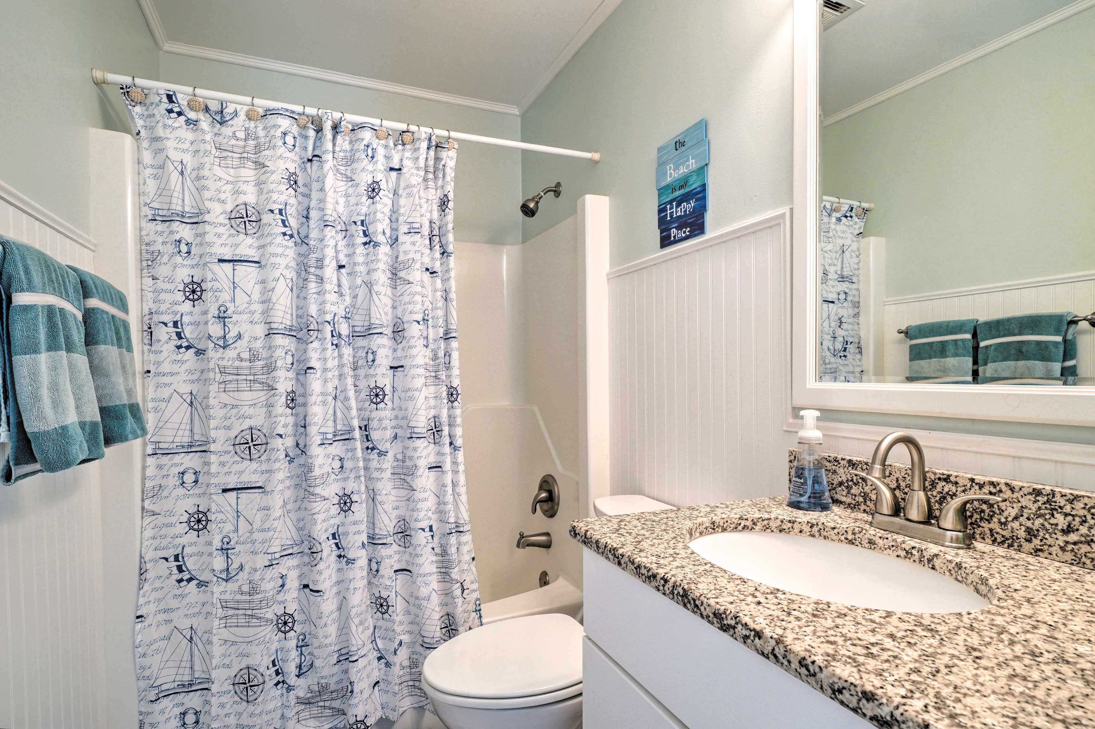 Een badkamer met een douchegordijn met nautisch thema, een wit toilet, een wastafel met granieten blad en een spiegel boven de wastafel.