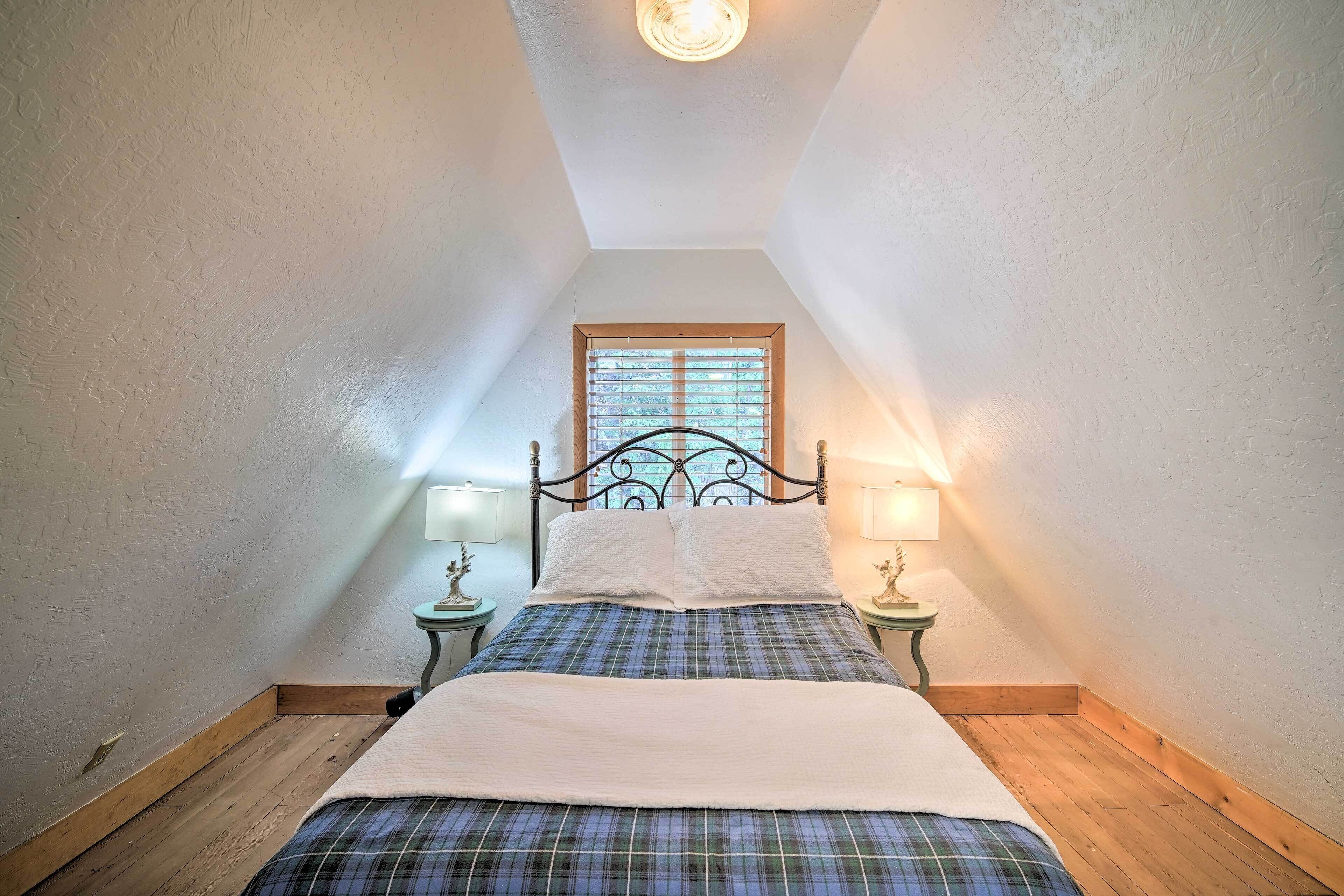 A bedroom with a sloped ceiling, a bed with a plaid bedspread, two bedside lamps, and a window with blinds.