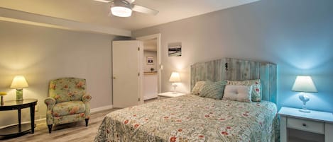 Un dormitorio con cama, una silla, una mesita de noche, un ventilador de techo y una puerta que da a otra habitación.