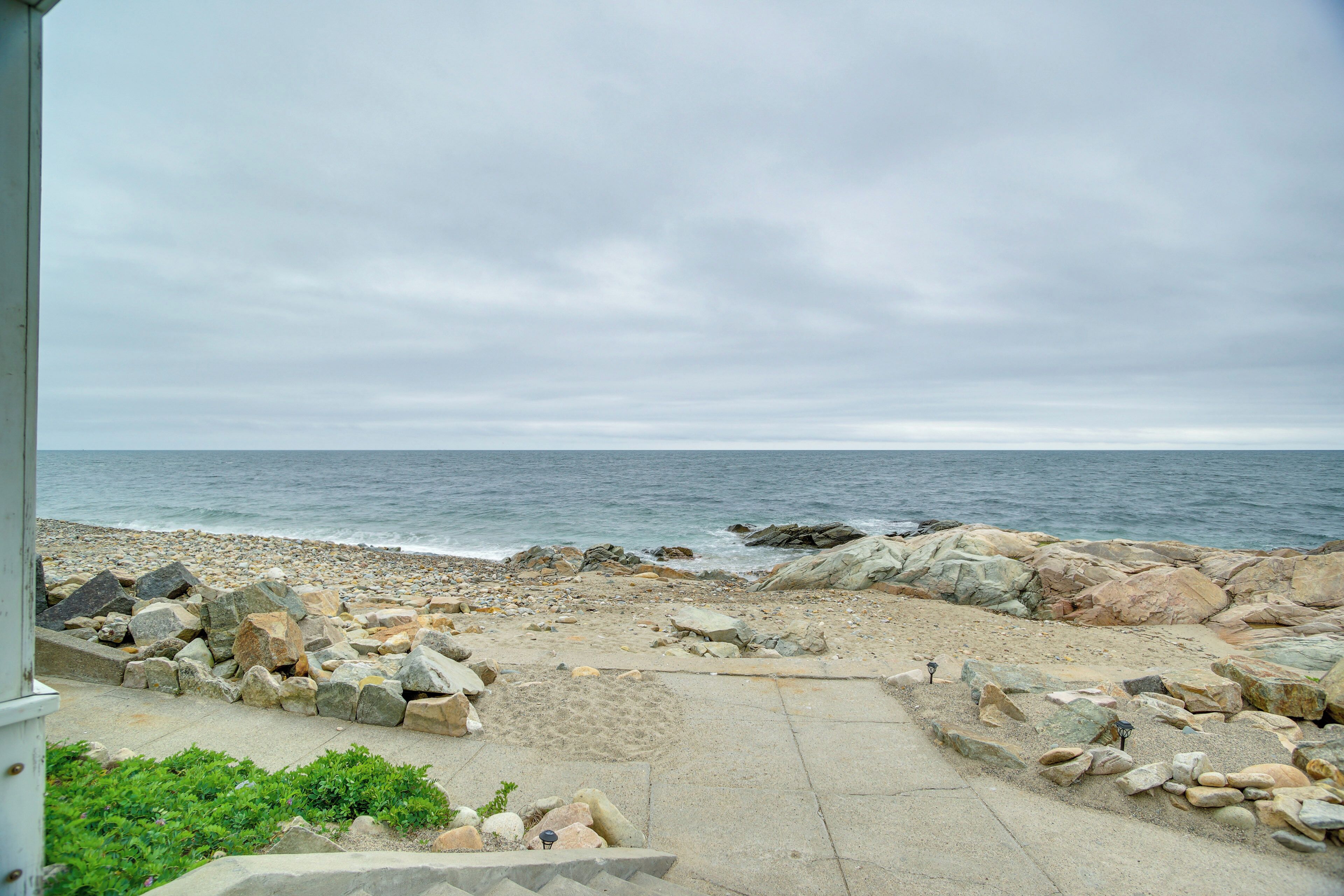 Une plage de galets avec un chemin de béton menant à la mer, sous un ciel nuageux.