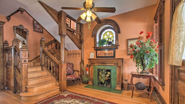 Una imponente escalera con barandas de madera y un candelabro, que conduce a una sala con chimenea y un arreglo floral.
