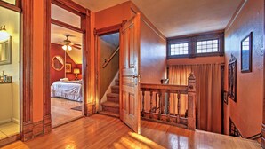 Una escalera de madera conduce a un dormitorio con una cama, un ventilador de techo y una ventana con cortinas.