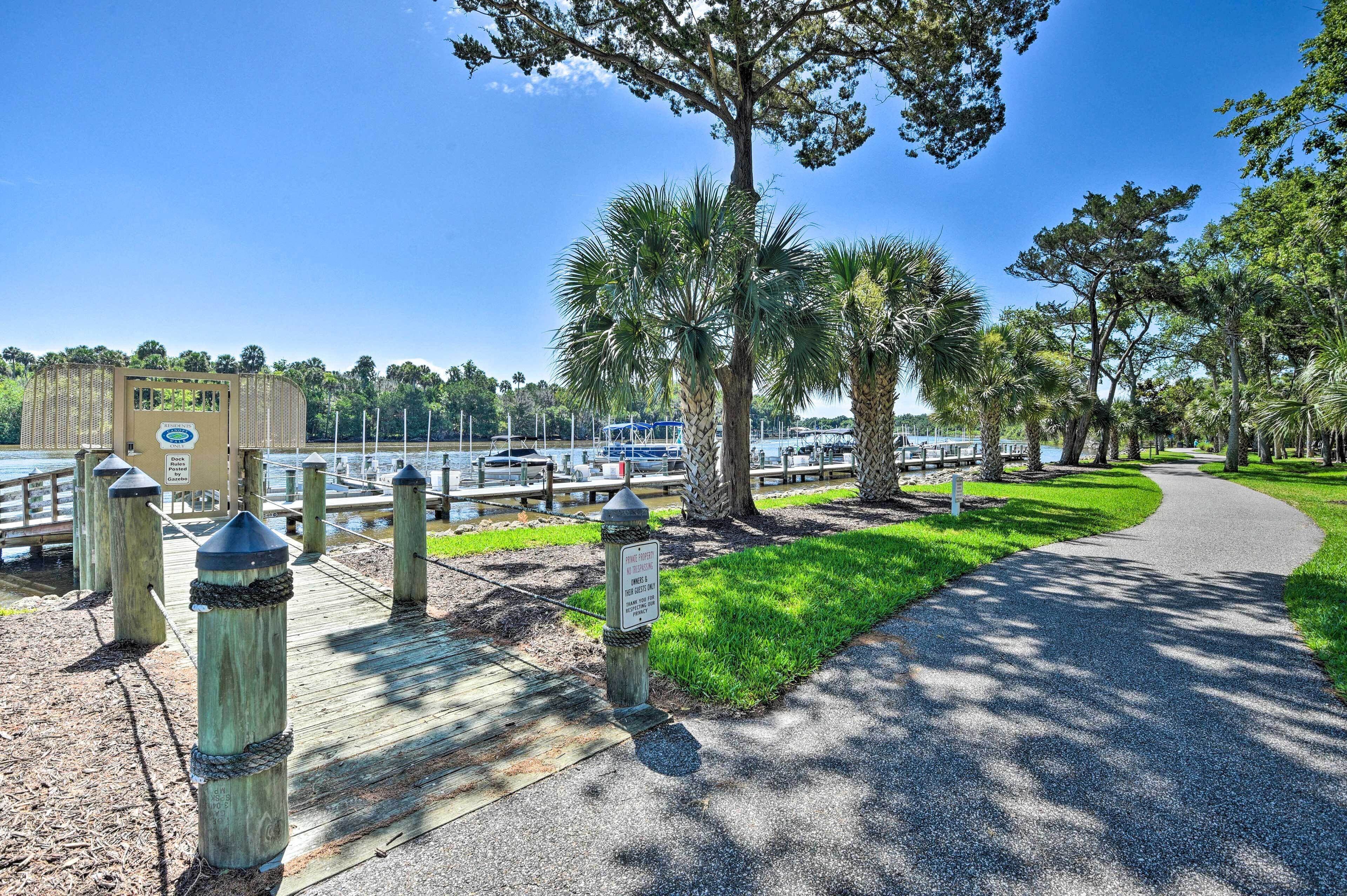 A wooden boardwalk with a paved path, surrounded by palm trees and a body of water with boats.