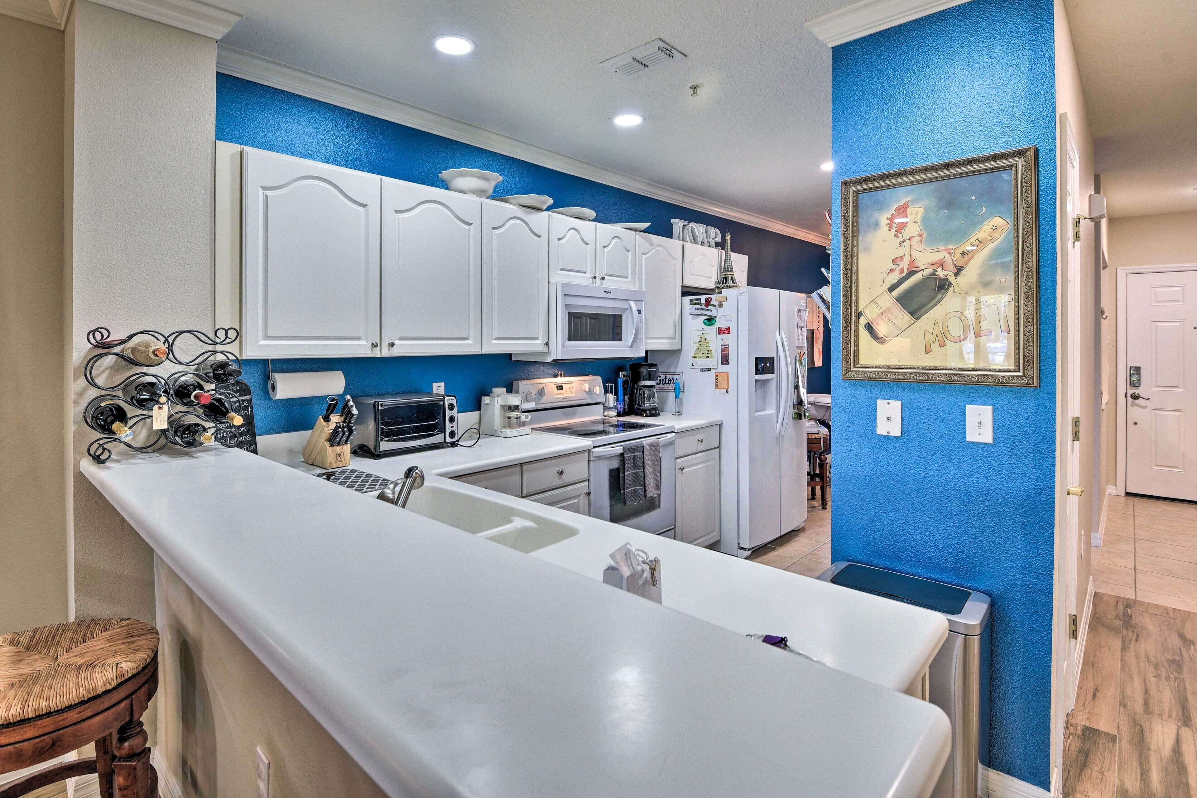 A modern kitchen with white cabinets, a microwave, and a refrigerator. There is a blue accent wall and a framed picture of a champagne bottle.