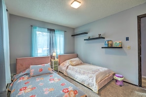 Um quarto com duas camas, uma janela com cortinas, uma estante com livros e um pequeno recipiente roxo no chão.