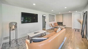 A modern living room with a leather sofa, a coffee table, and a floor lamp. There is a fireplace and a door in the background. - Artsy Upstate Getaway w/ Huge Private Yard! (Whitesboro)