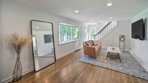 A spacious living room with hardwood flooring, a large mirror, a beige sofa, and a staircase leading to another level. - Artsy Upstate Getaway w/ Huge Private Yard! (Whitesboro)