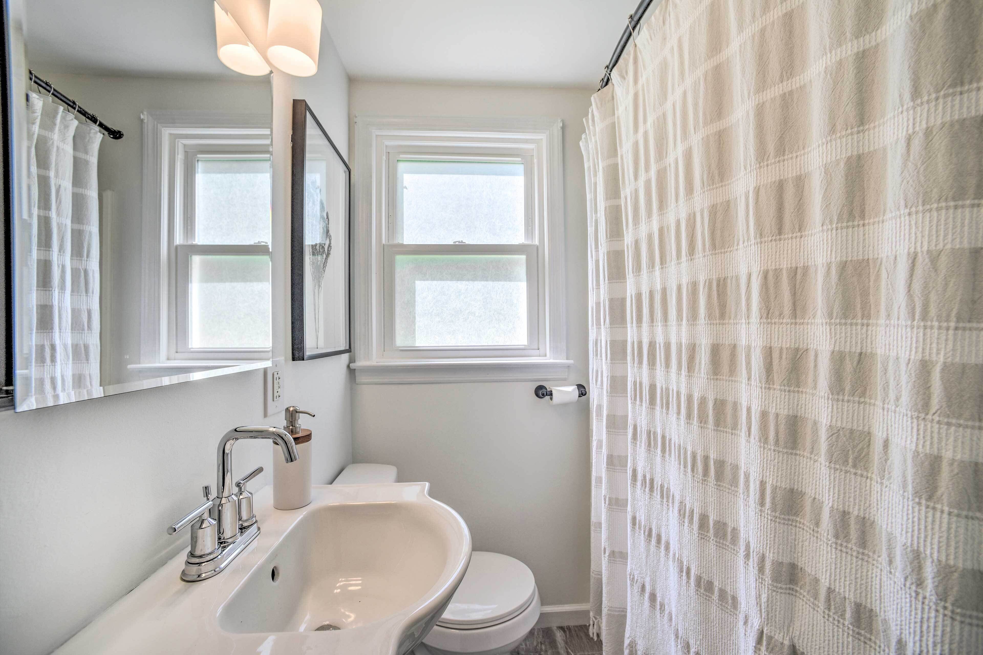 A bathroom with a white sink, mirror, and toilet. There are two windows above the sink and toilet, and a shower curtain.