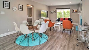 A modern living room with a glass dining table, white chairs, an orange sofa, and a TV on a white stand. - Palm Springs Retreat w/ Private Pool & Spa! (Palm Springs)