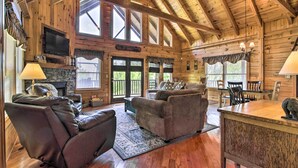 Un intérieur spacieux de chalet en bois, doté d’une cheminée, de canapés en cuir, d’un coin repas et de grandes fenêtres.