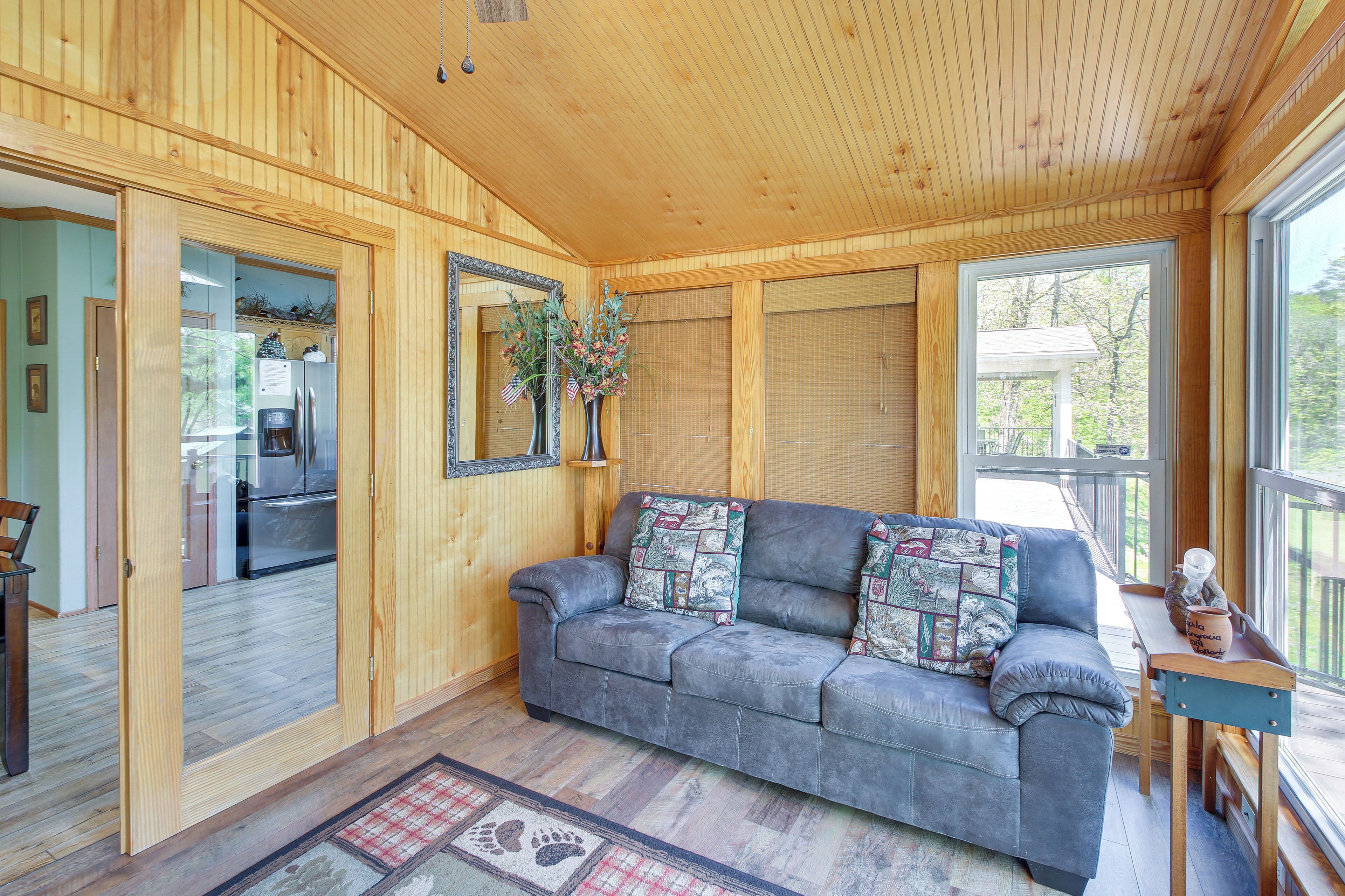 A cozy living room with a grey sofa, wooden walls, a large window, and a wooden ceiling.