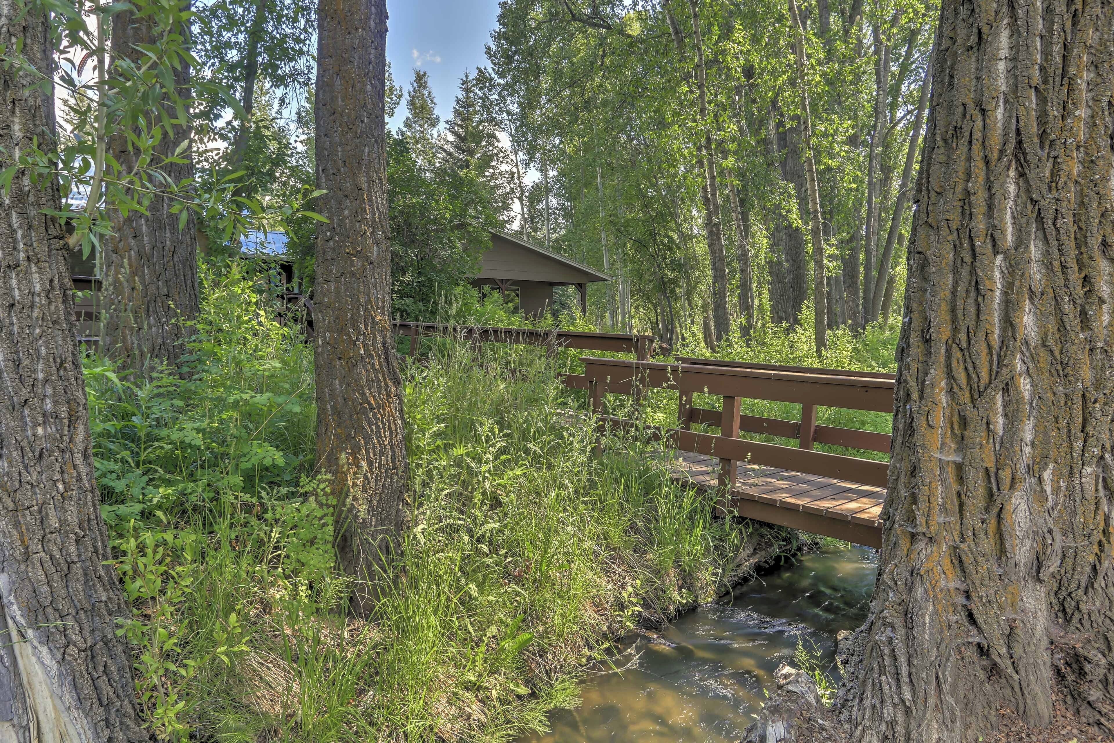 Eine Holzbrücke über einen Bach in bewaldeter Landschaft, im Hintergrund ein kleines Gebäude.