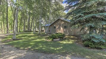 Une cabane dotée d’un toit en pente, entourée d’une zone boisée avec de grands arbres et une pelouse bien entretenue.