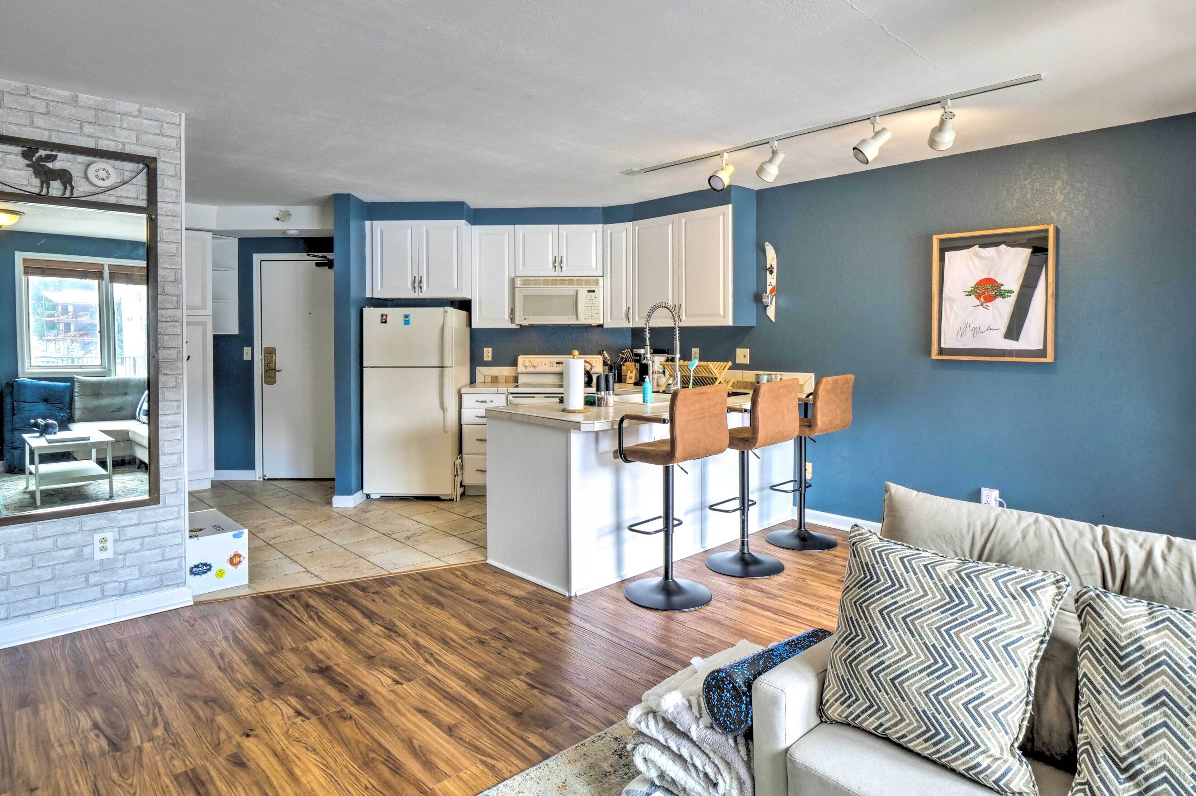 A modern living space with a kitchen, a dining area, and a seating area with a sofa and bar stools.