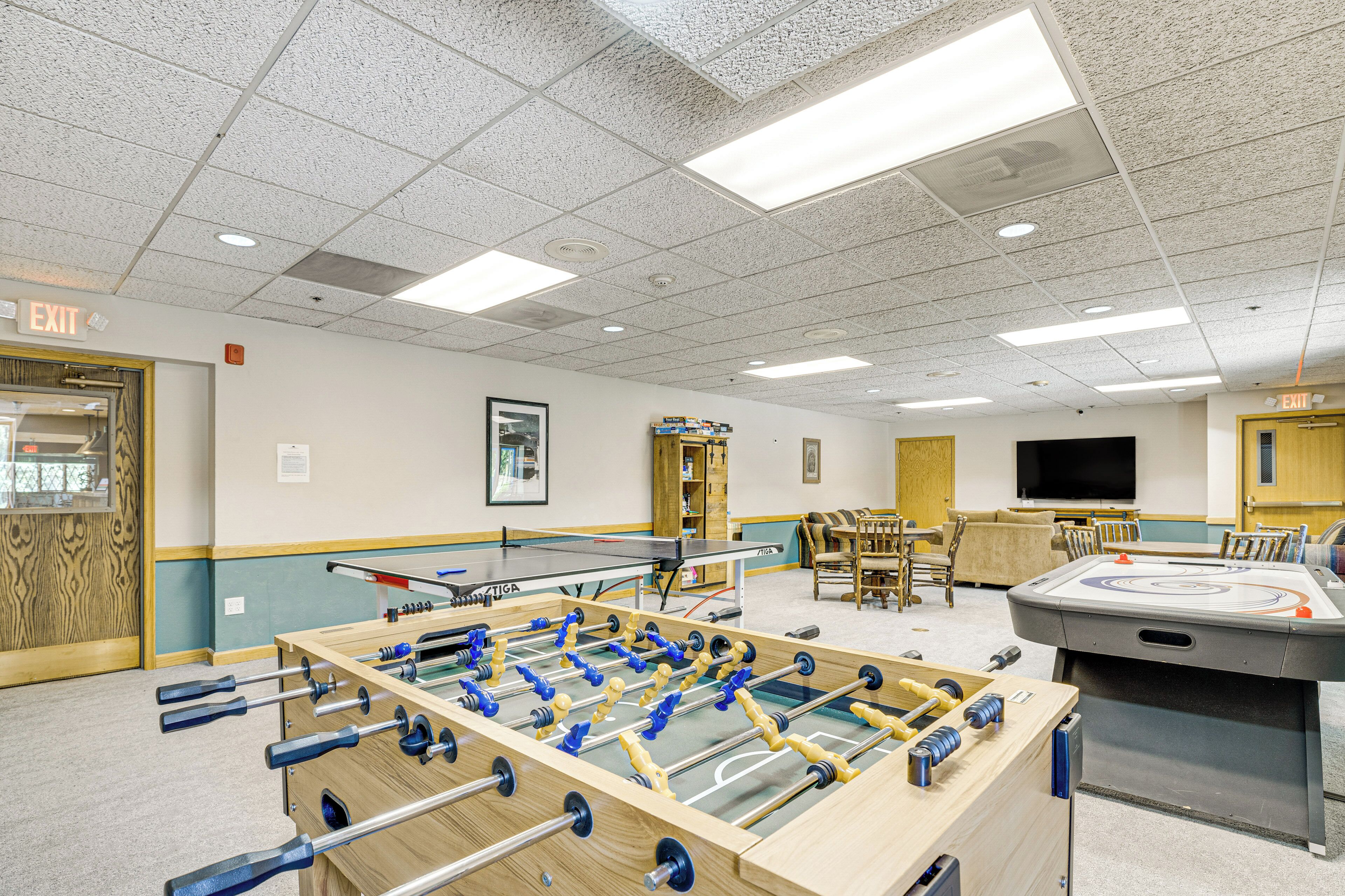 A room with a foosball table, a ping pong table, and a shuffleboard table. There are chairs and a television in the room.