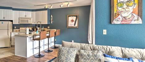 Una cocina moderna con armarios blancos, un microondas y una zona de bar con taburetes. Hay un sofá con cojines estampados y un cuadro en la pared.