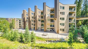 A multi-story residential building with balconies, a covered outdoor area, and a natural surrounding with trees and grass.