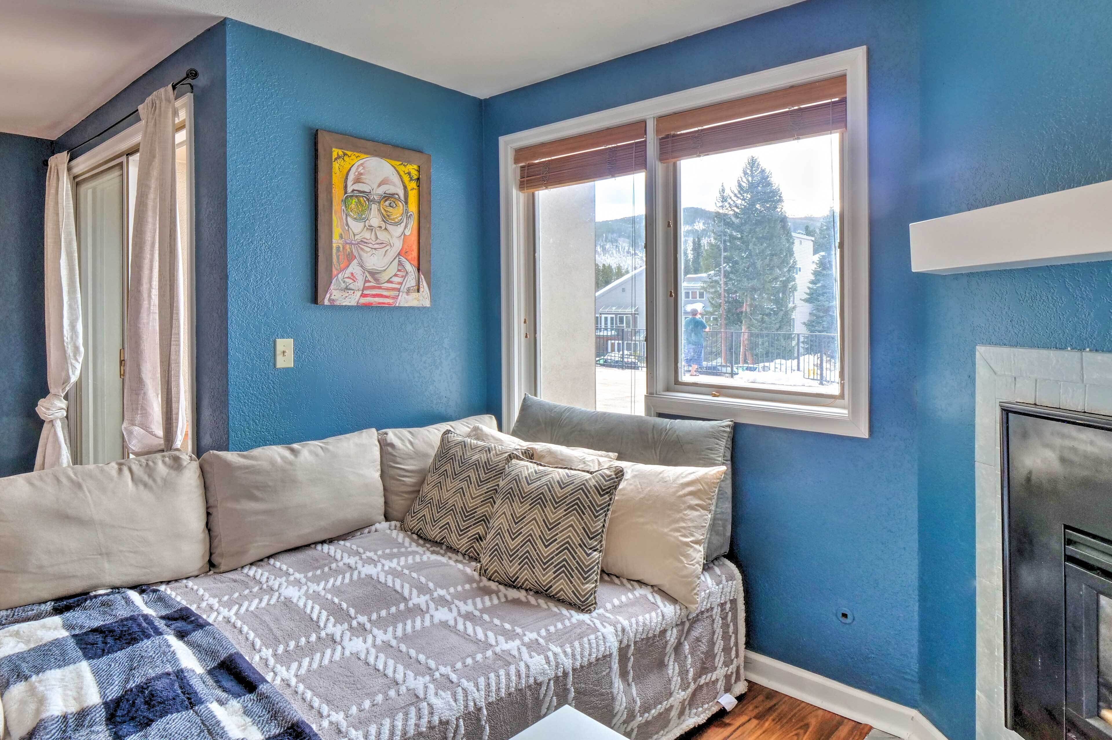 Une chambre avec un mur bleu, une fenêtre avec des stores, un lit avec une literie à motifs, un tableau au mur et une cheminée.