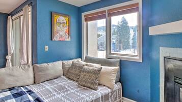 Une chambre avec un mur bleu, une fenêtre avec des stores, un lit avec une literie à motifs, un tableau au mur et une cheminée.
