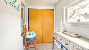 A hallway with a wooden door, a window with a view of houses, a chair with a colorful bag, and a wooden cabinet with drawers. - 'ania's Retreat' w/ Deck: Walk to Lake Michigan! (Two Rivers)