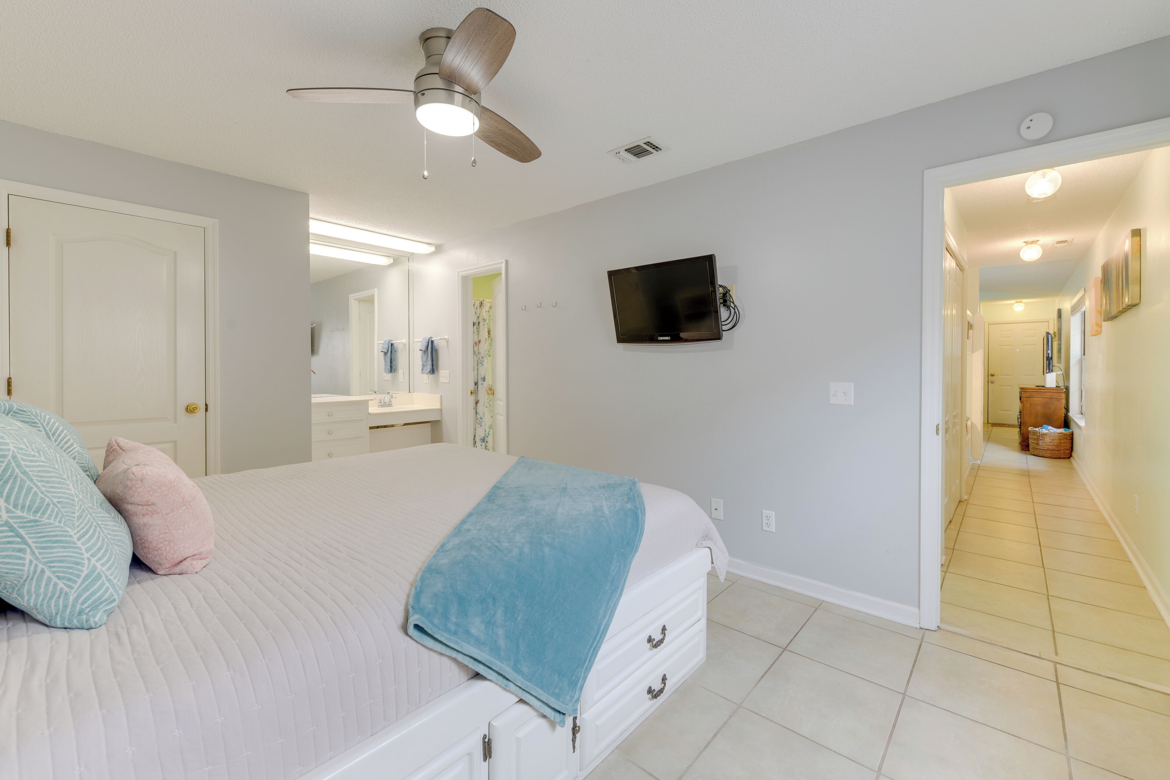 A bedroom with a bed, a ceiling fan, a TV, and a hallway leading to another room.