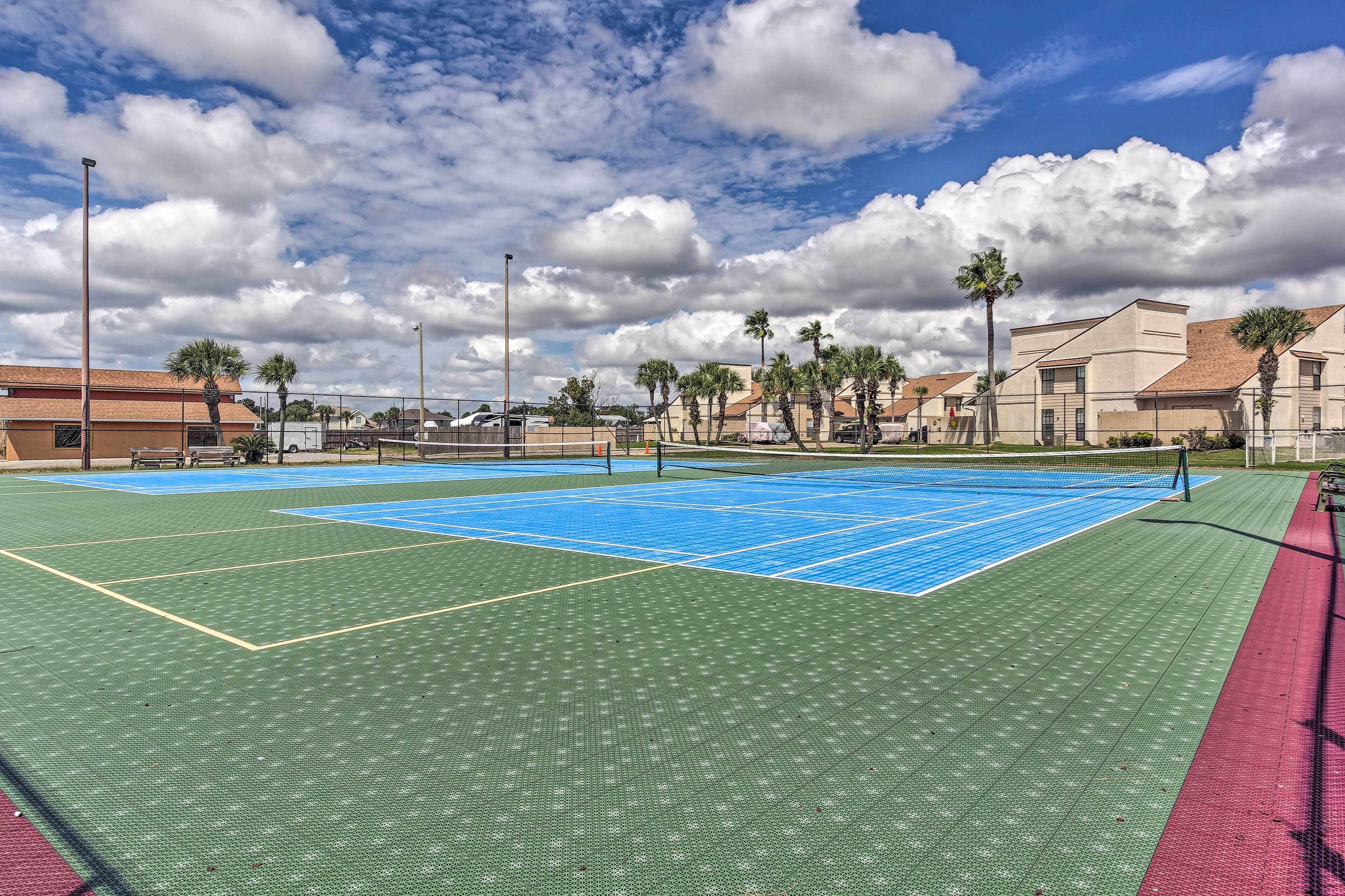 A tennis court with a blue and green surface, surrounded by palm trees and buildings.