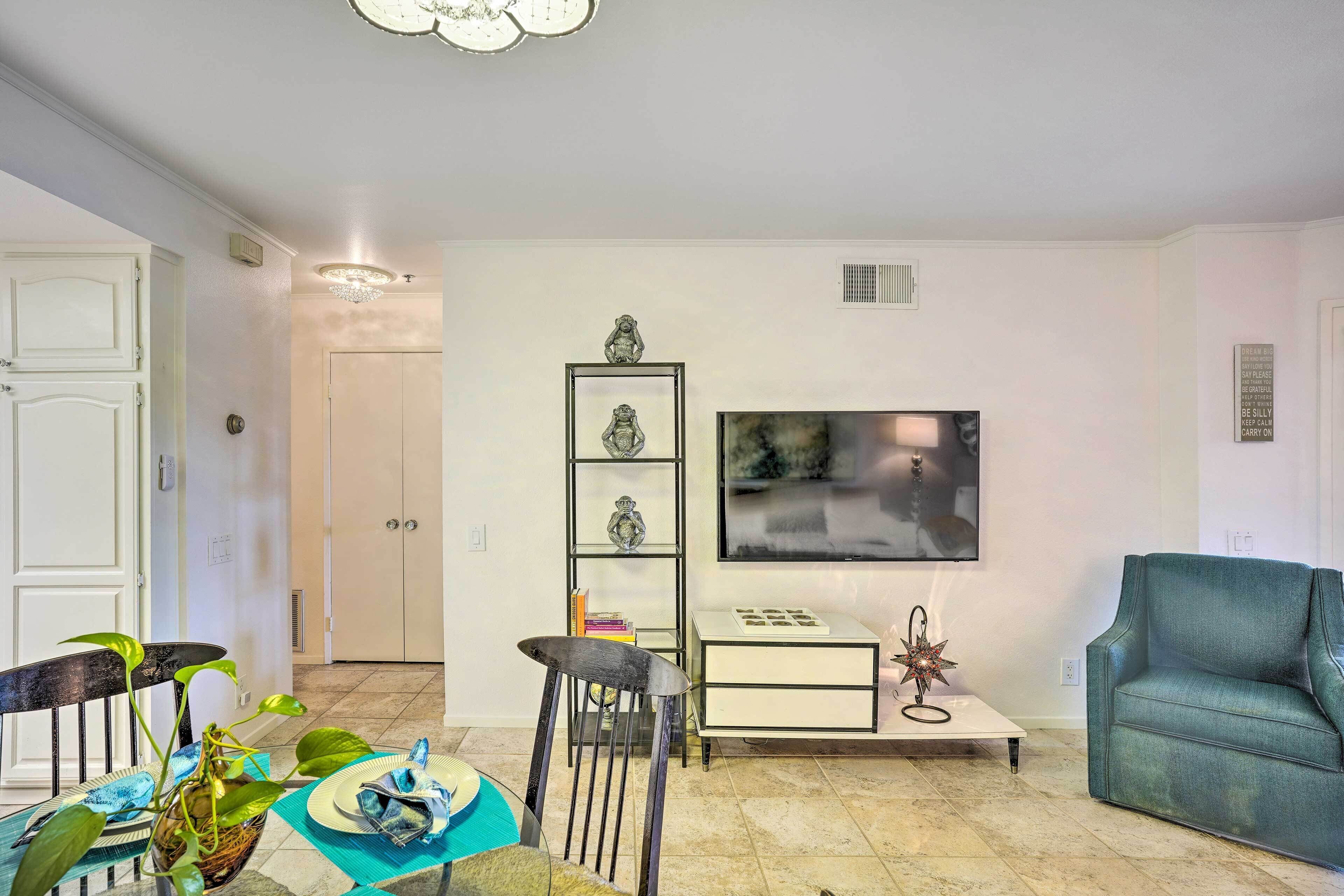 A living room with a teal sofa, a dining table with chairs, a TV mounted on the wall, and a bookshelf.