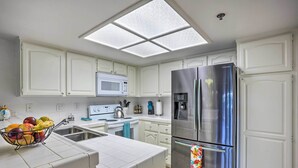 A modern kitchen with white cabinetry, a stainless steel refrigerator, a microwave, and a fruit basket on the counter. - Condo w/ Patio, Walk to Downtown Palm Springs! (Palm Springs)