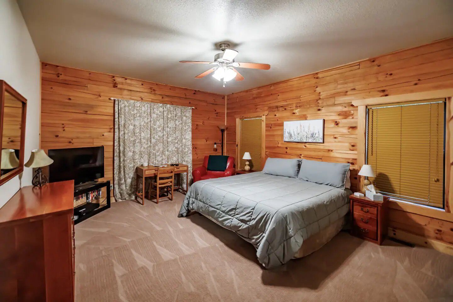 Ein Schlafzimmer mit Holzwänden, ausgestattet mit einem Bett, Schreibtisch, Stuhl, Fernseher und Deckenventilator.