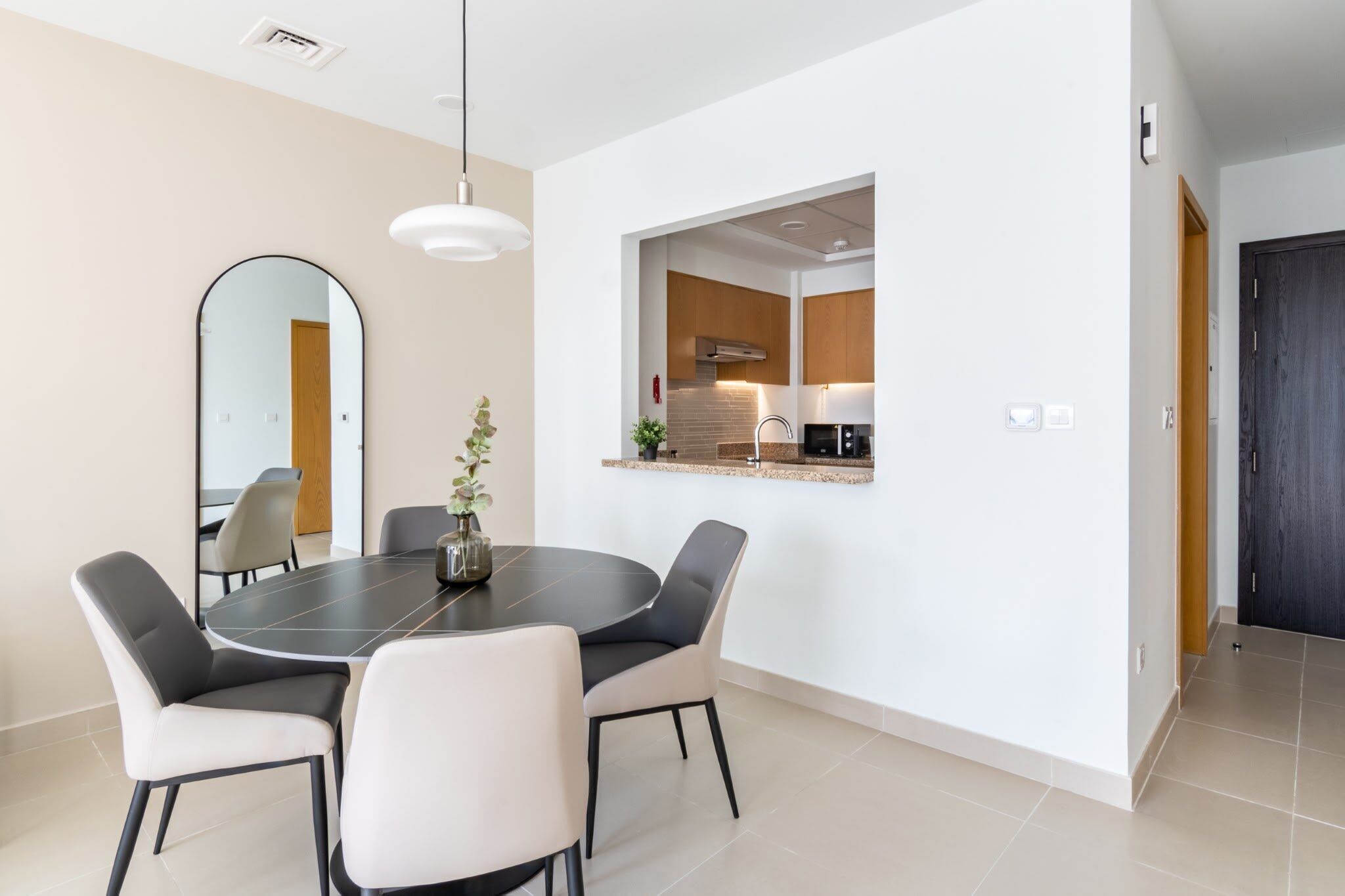 A modern dining area with a round table and chairs, a kitchen visible through an open archway, and a dark door to the right.