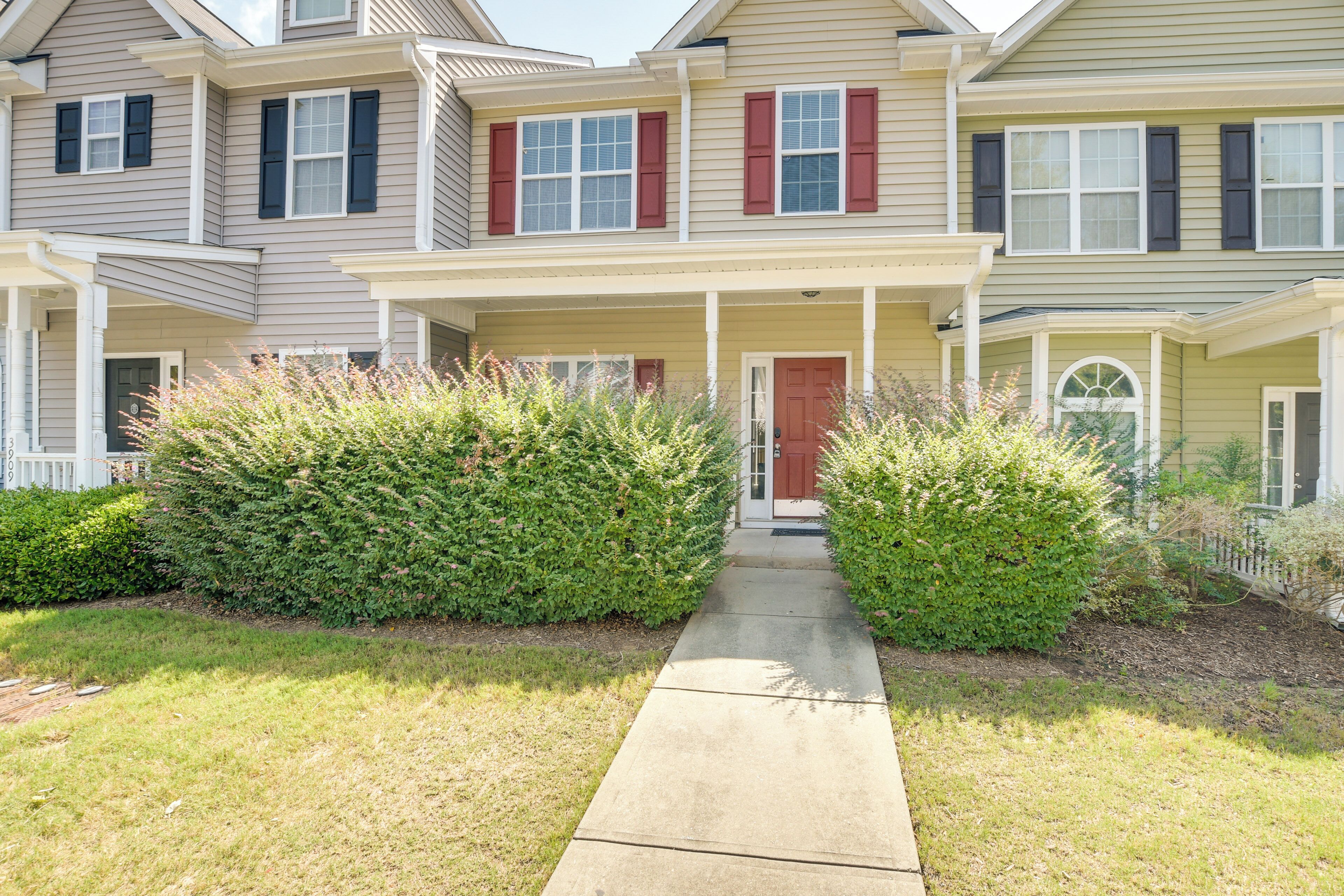 A two-story residential building with a front porch, a red door, and a pathway leading to the entrance.