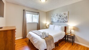 A neatly made bed with a white comforter and a beige throw blanket. There is a wooden dresser to the left of the bed. A painting hangs above the bed, and a small table with a lamp is on the right side of the bed. - Lovely Colorado Springs Home: Mtn Views & Hot Tub! (Colorado Springs)