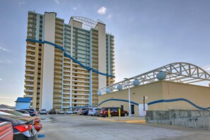 A high-rise hotel with multiple balconies, a parking area with several cars, and a distinctive curved architectural feature. - Oceanfront Panama City Beach Condo w/ Pool Access! (Panama City Beach)