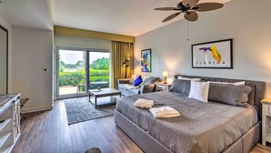 Quarto de hotel moderno com uma cama grande, ventoinha no teto, sofá, escrivaninha e vista para o verde lá fora.