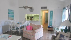 A living room with a striped sofa, white chairs, a dining table, and a kitchen in the background.