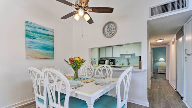 A dining area with a white table and chairs, a ceiling fan, a clock, and a kitchen in the background.