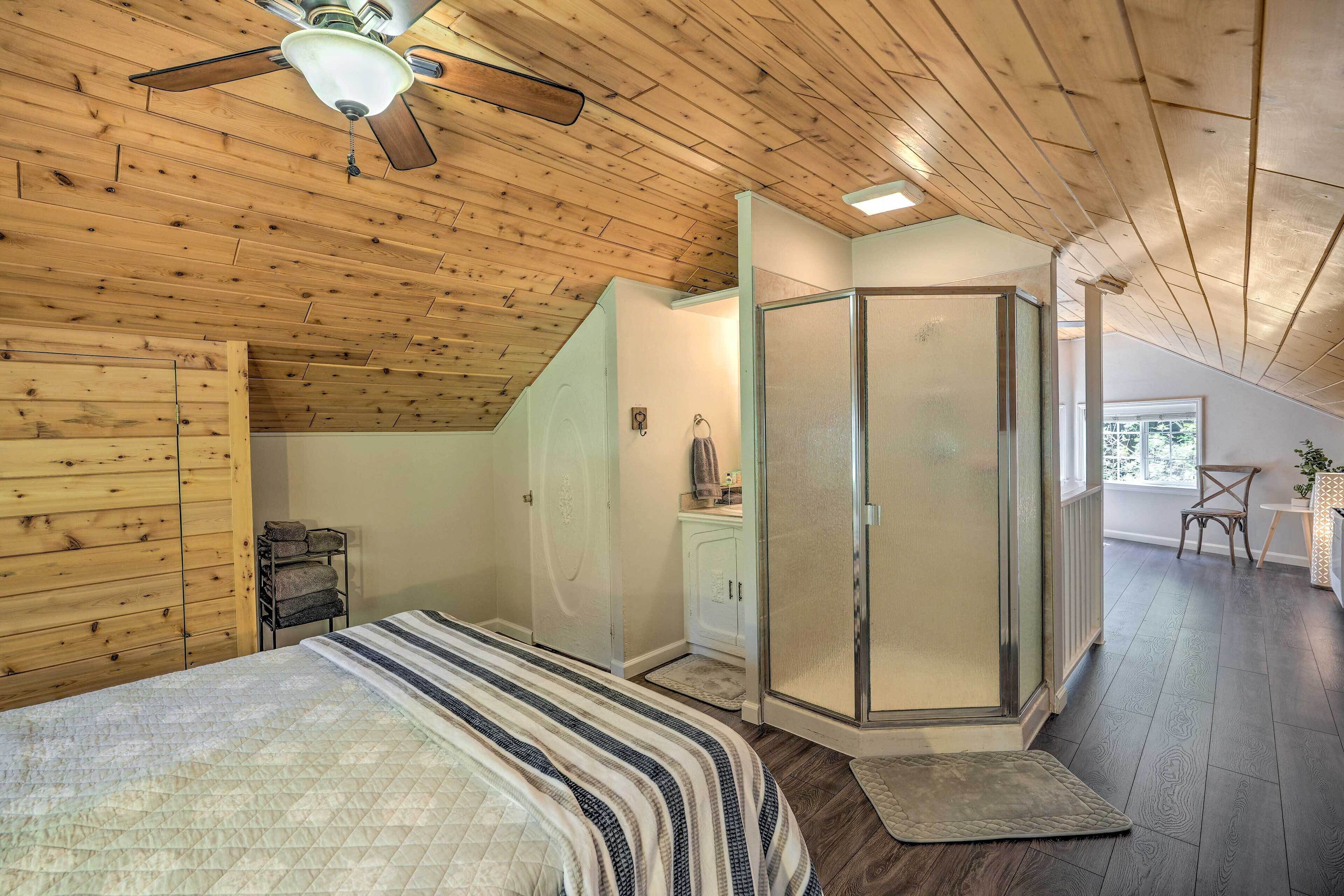 A bedroom with a wooden ceiling, a bed with a striped blanket, a walk-in closet, a shower with frosted glass doors, and a view of the outdoors.