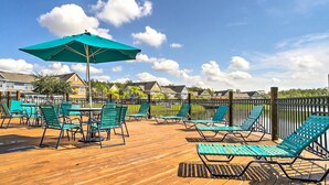 Une terrasse en bois avec des fauteuils de jardin verts et un grand parasol, donnant sur une étendue d’eau avec des maisons en arrière-plan.