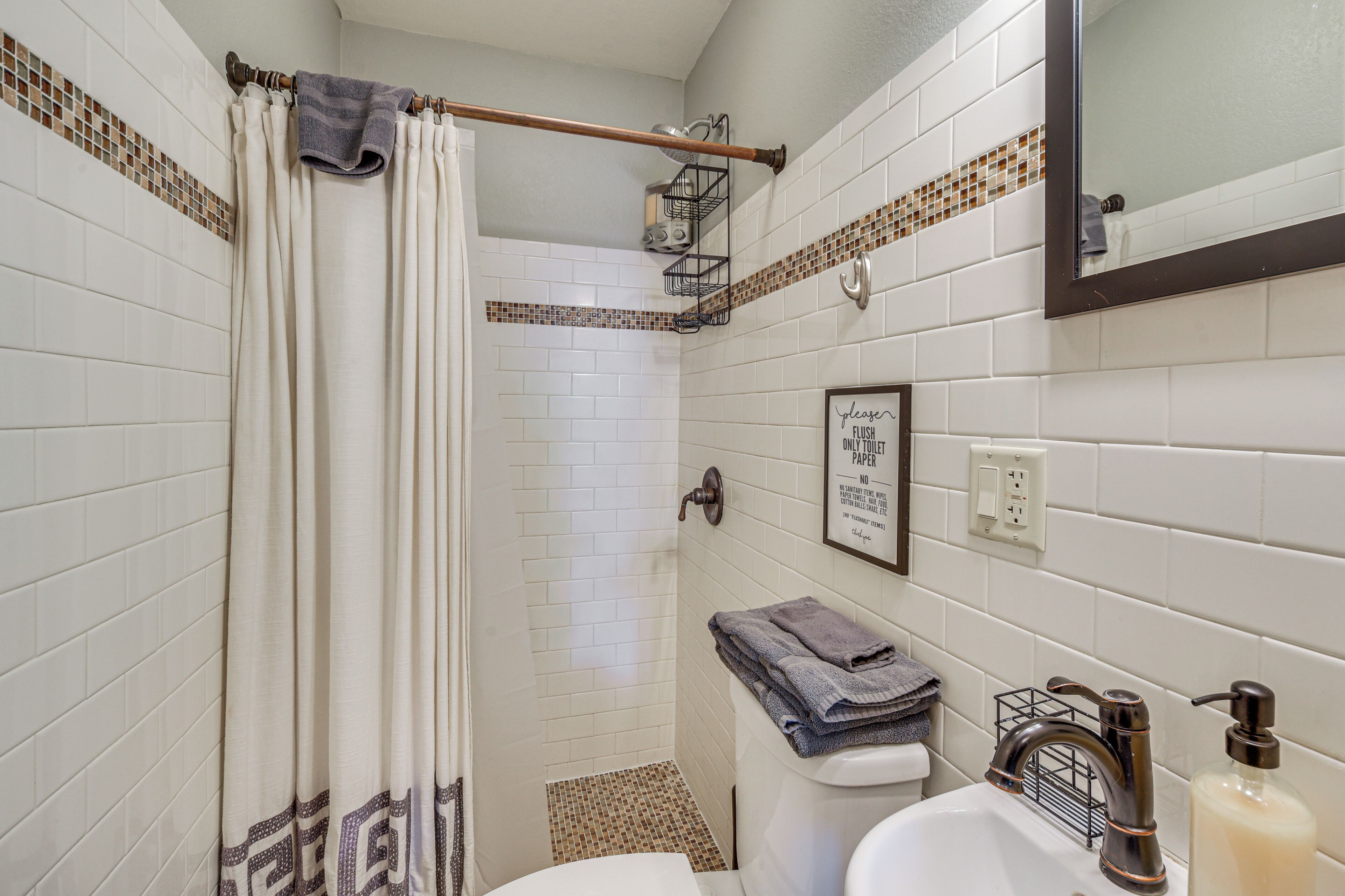 A bathroom with white tiled walls, a shower curtain, a toilet, a sink with a soap dispenser, and a mirror.