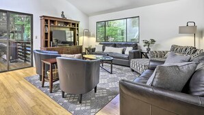 A modern living room with a sofa, armchairs, a coffee table, and a TV. There is a bookshelf and a large window providing a view of the outdoors. - Tranquil Guerneville Home w/ Redwood Views (Guerneville)