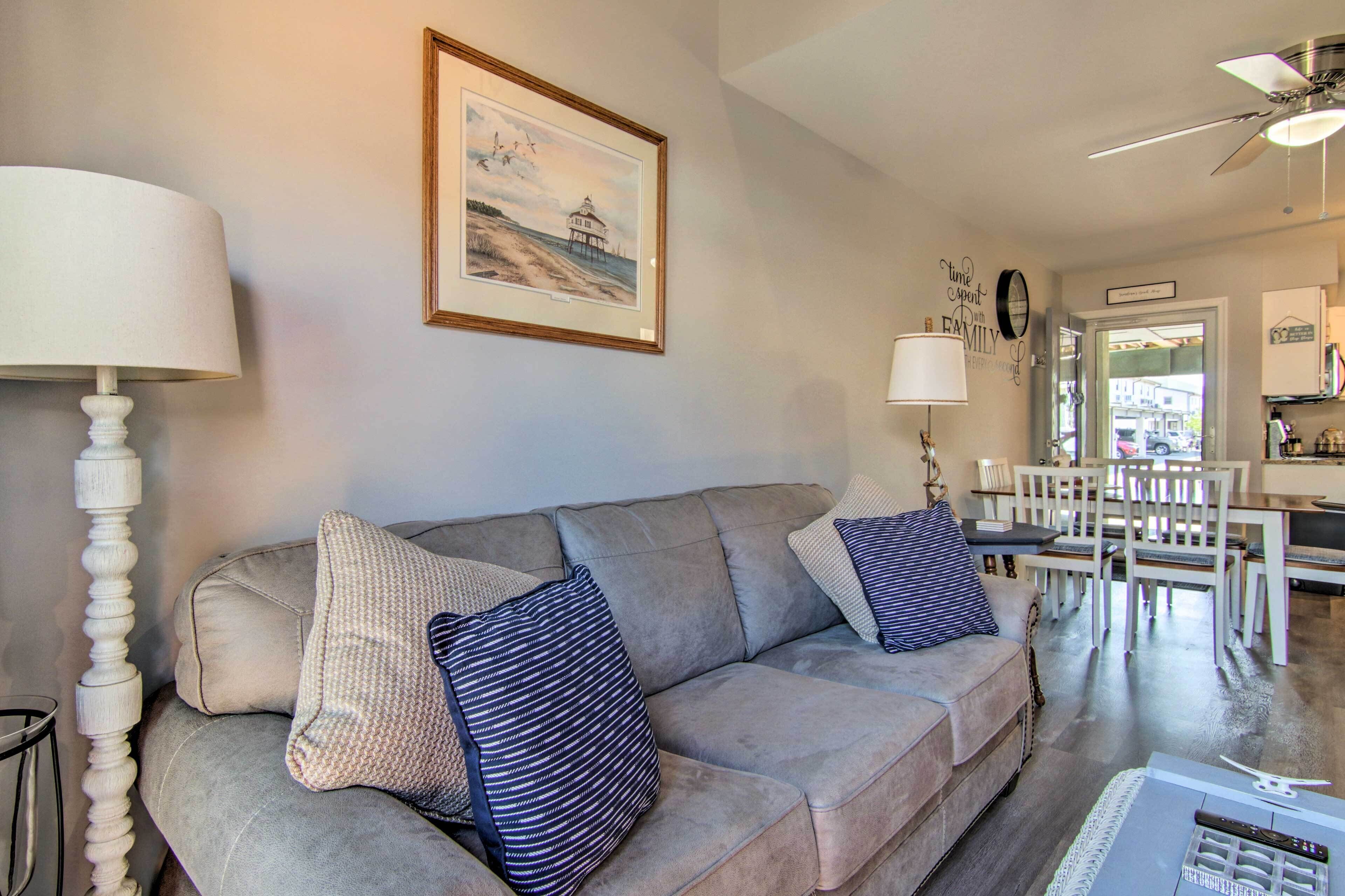 A living room with a grey sofa, two pillows, a framed picture on the wall, a lamp, and a ceiling fan.