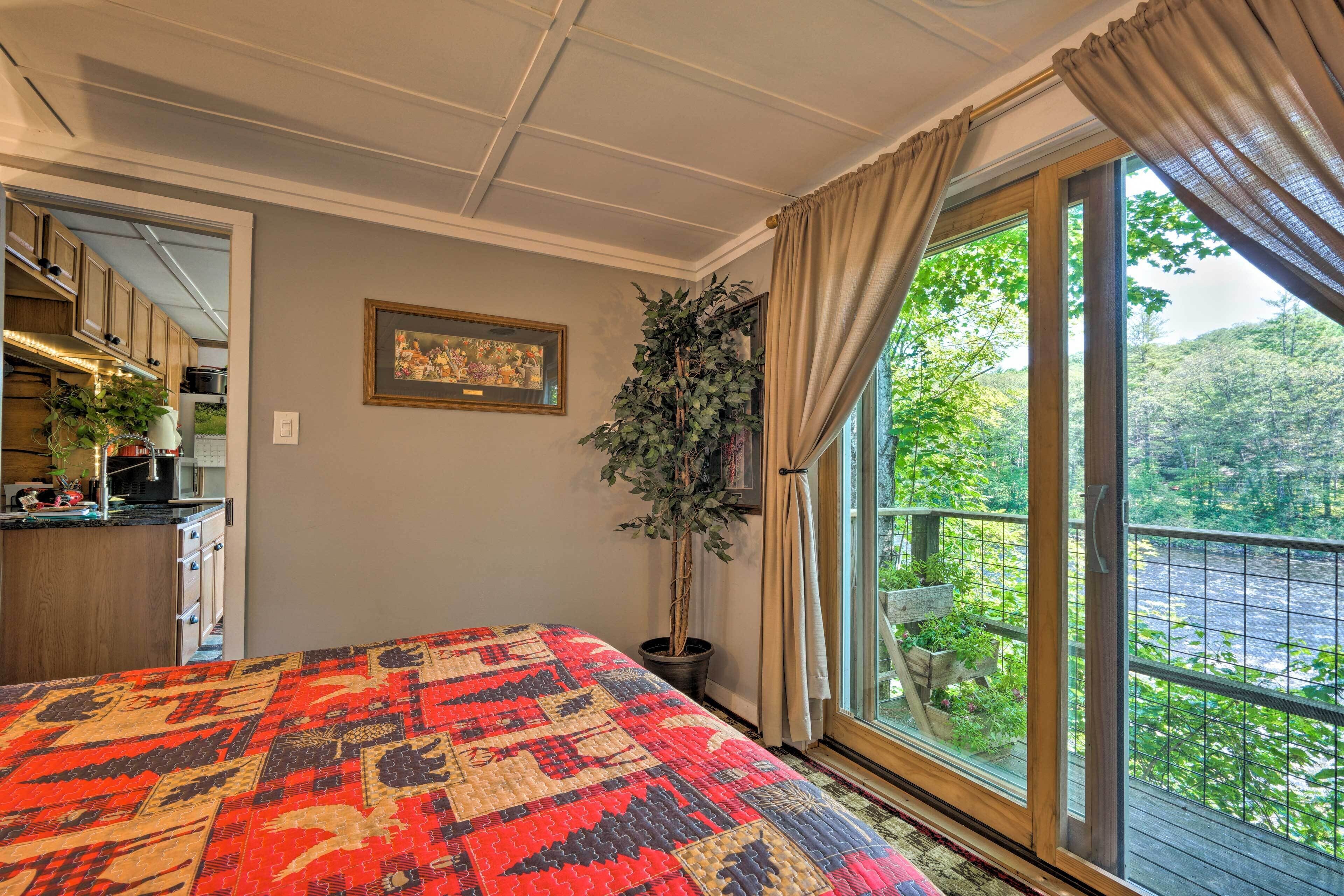 A bedroom with a large bed, a potted plant, a window with a view of greenery, and a kitchen area visible through an open door.
