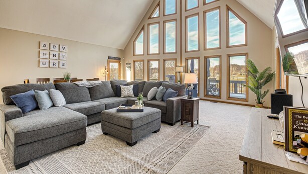 A spacious living room with a large sectional sofa, a coffee table, and a wooden side table. There are multiple windows providing natural light and a view of the outdoors. - Apple Valley Home, Shared Golf Course On-site (Howard)