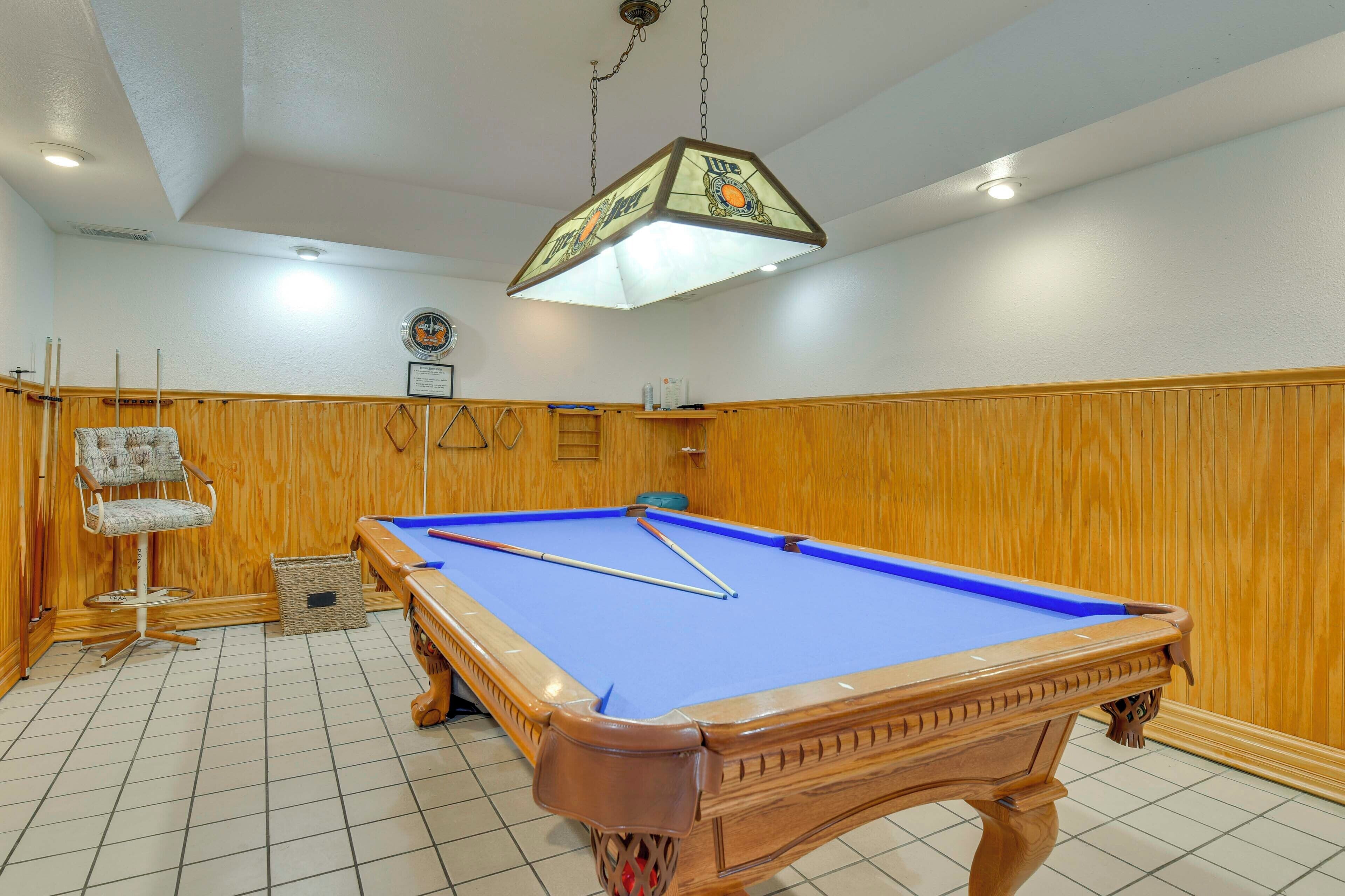 Una mesa de billar con estructura de madera, una lámpara colgante, una silla y una pequeña cesta en el suelo.