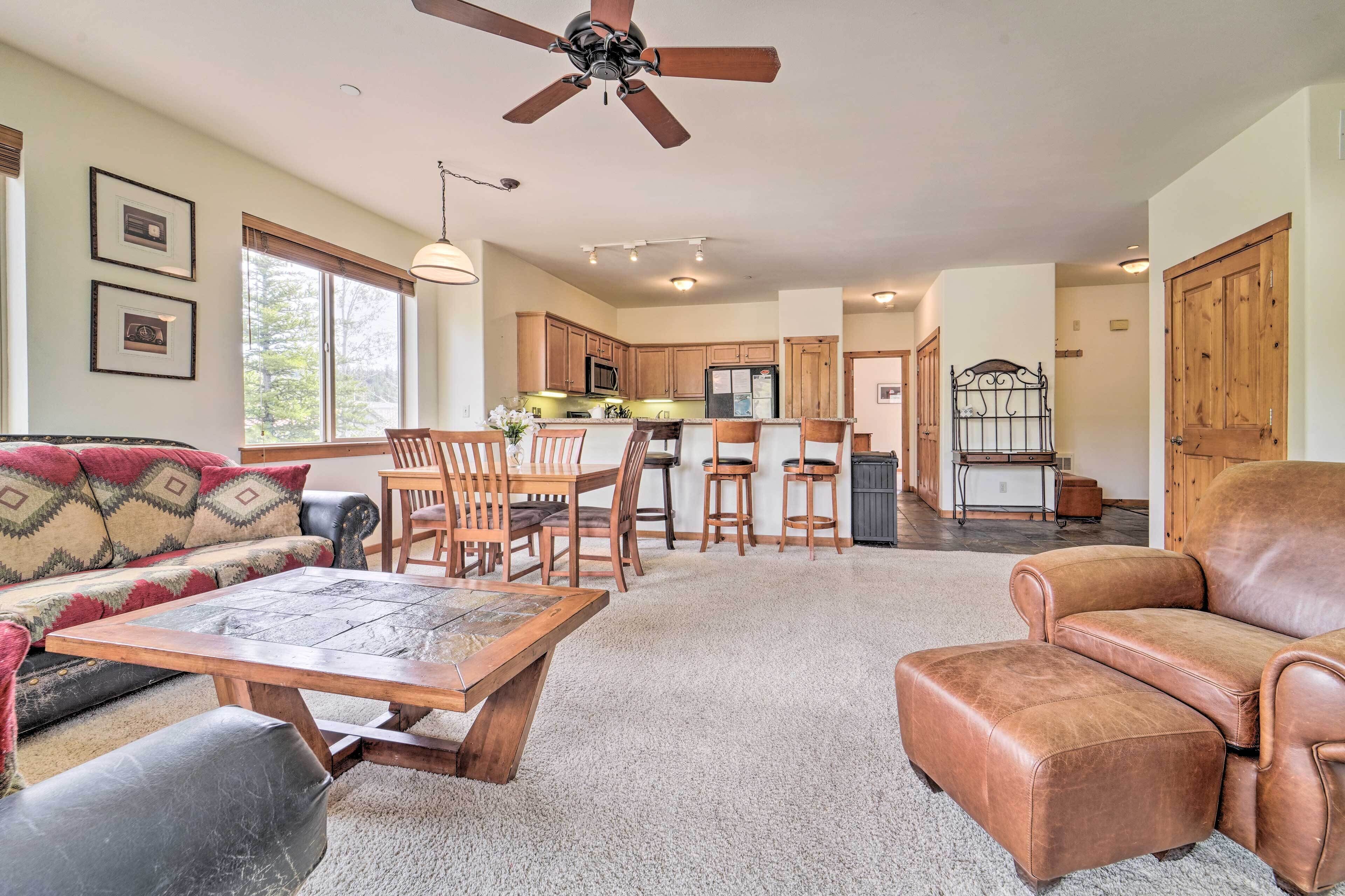 A spacious living area with a sofa, a wooden coffee table, and a leather armchair. A kitchen with wooden cabinets and a dining area with chairs is visible in the background.