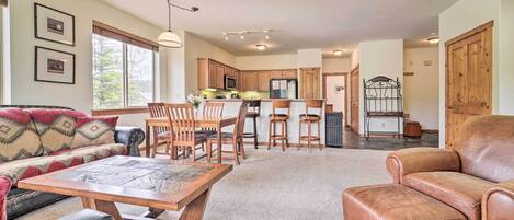 A spacious living area with a sofa, a wooden coffee table, and a leather armchair. A kitchen with wooden cabinets and a dining area with chairs is visible in the background.