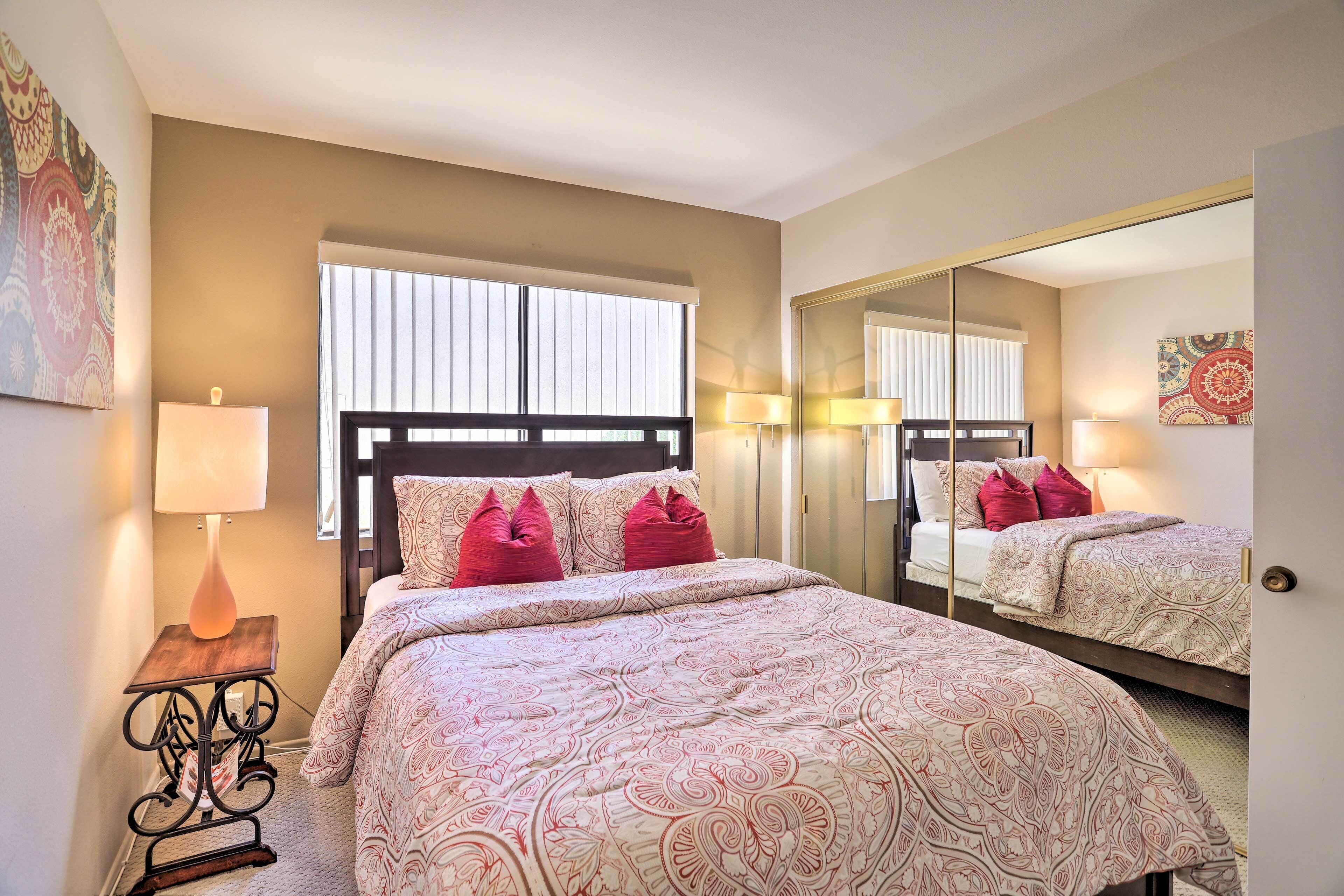 A bedroom with a large bed, a bedside table with a lamp, a window with blinds, and a mirror on the wall.