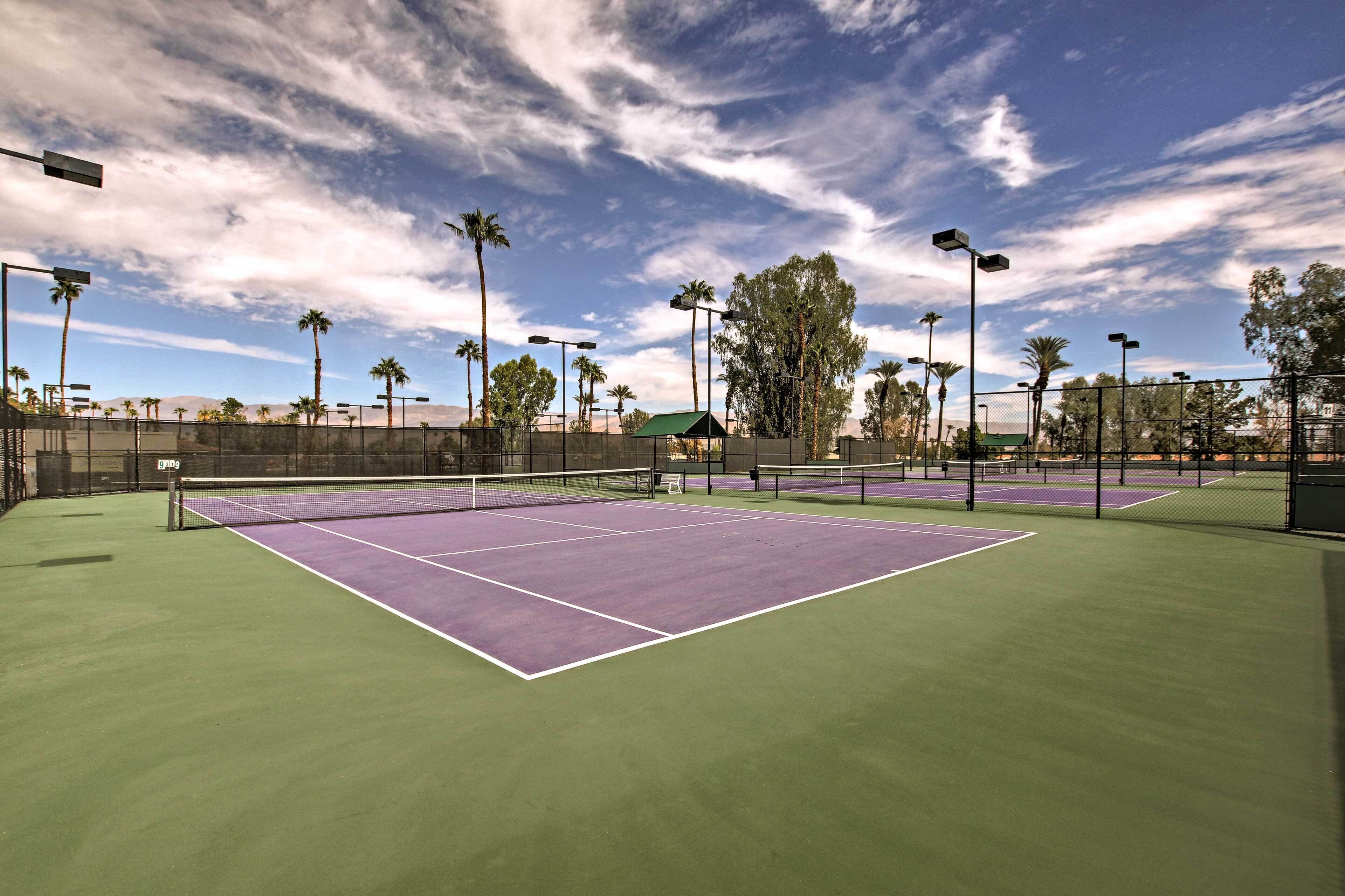 Een tennisbaan buiten met een groene ondergrond, omringd door een hek en palmbomen op de achtergrond.