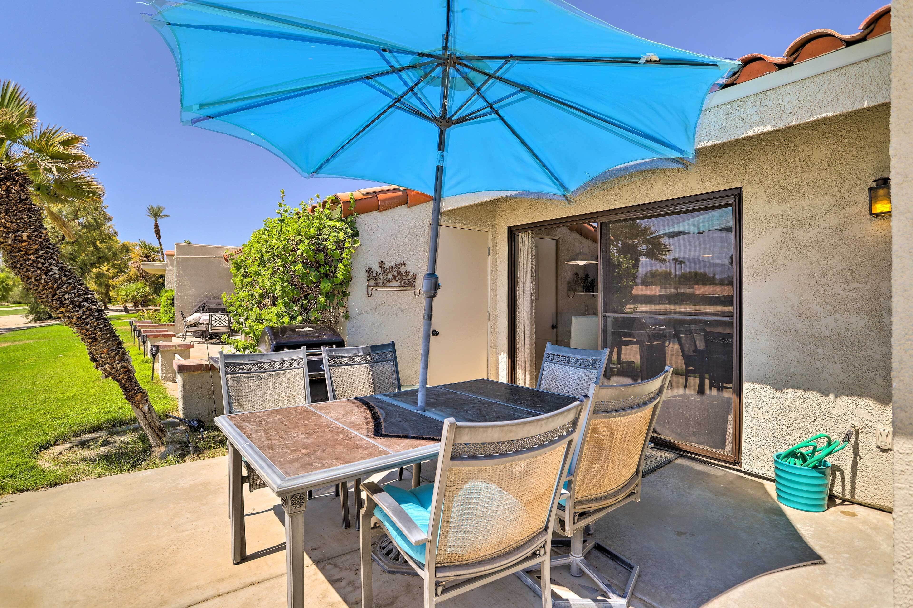An outdoor patio with a table and chairs, a blue umbrella, and a view of the surrounding area.